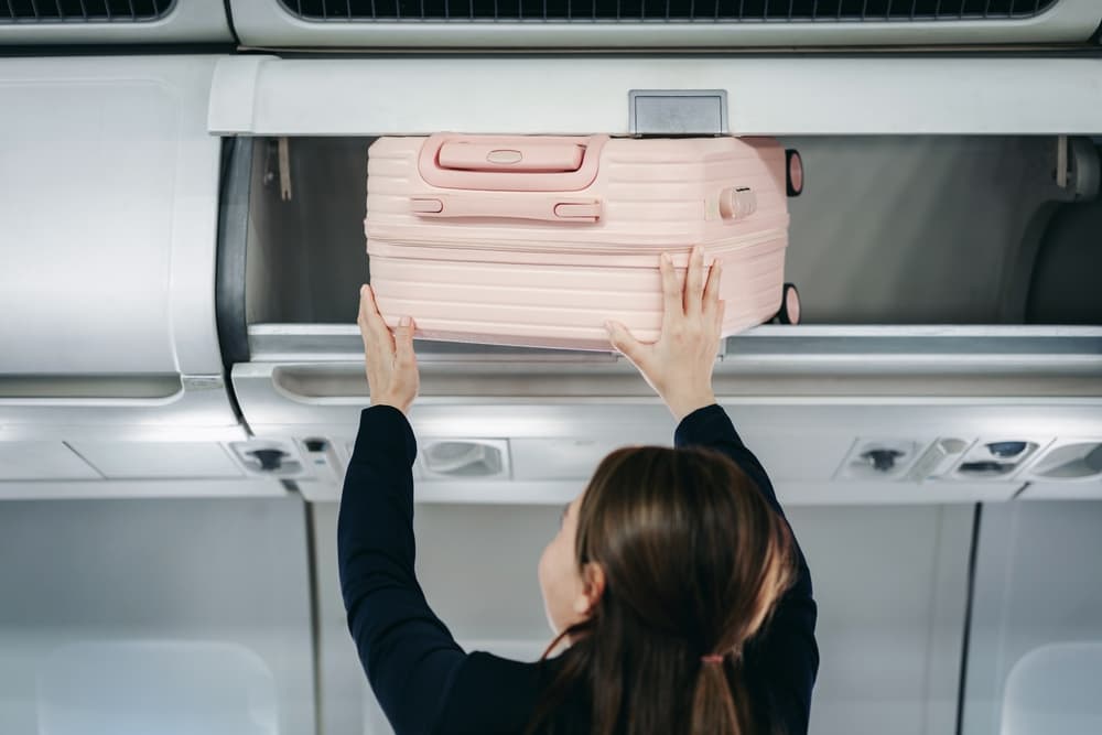 female traveler stowing pink luggage overhead bin on passenger airplane